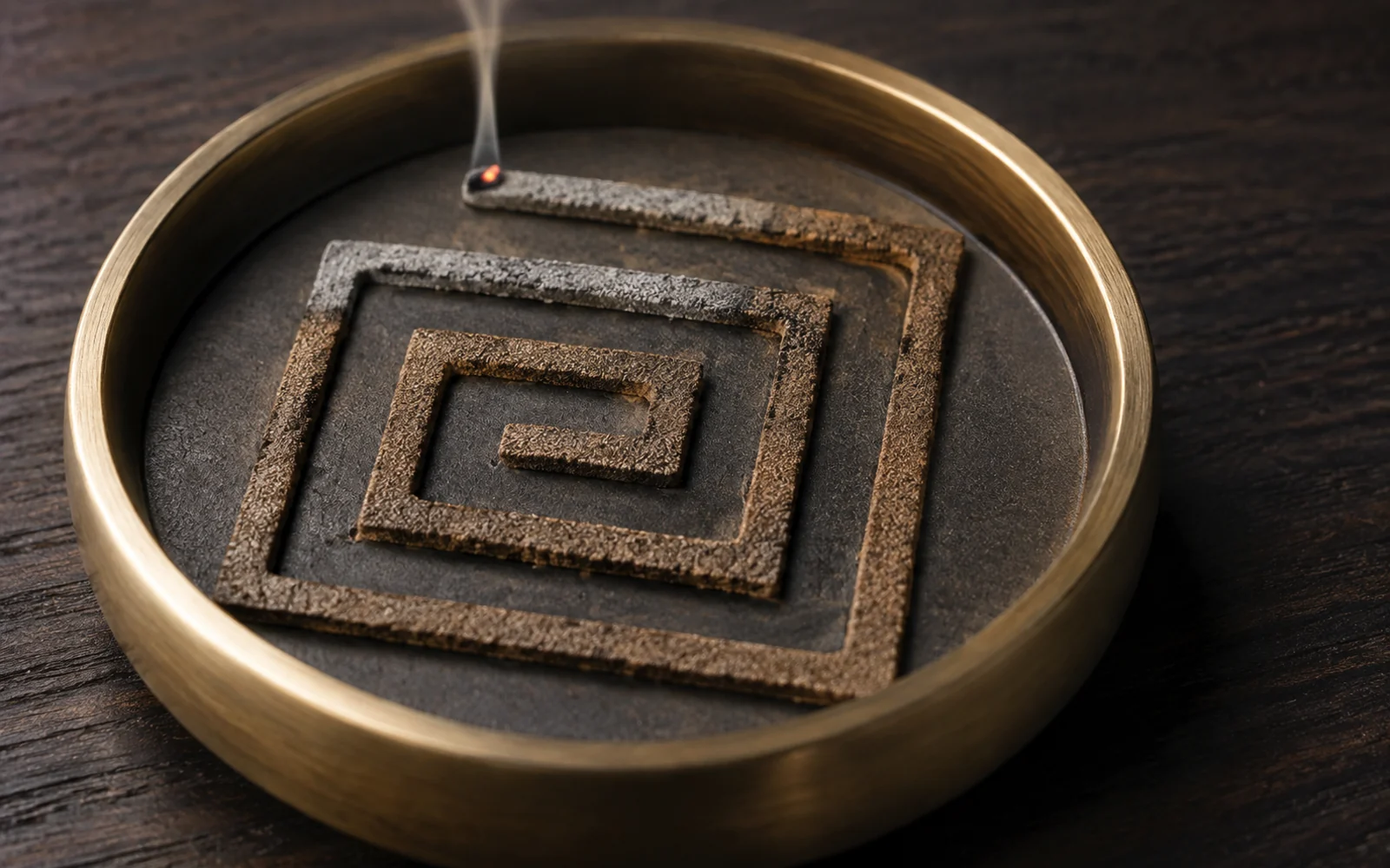 Seal incense powder pressed into a square-spiral 香篆 pattern in a brass censer, lit at one end with the burnt section fallen to grey ash